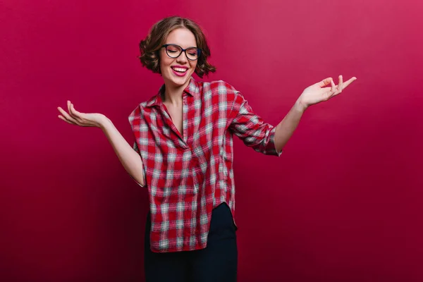 Happy girl wears checkered shirt and black pants posing with hands up. Studio portrait of relaxed white lady in glasses..