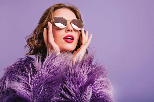 Close-up photo of inspired trendy lady in sparkle glasses looking up with mouth open. Indoor portrait of elegant young woman in purple fur coat..