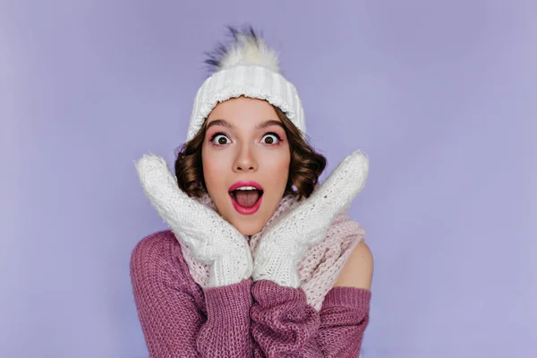 Shocked european woman wears soft white mittens posing in studio. Indoor portrait of short-haired female model wears woolen hat and gloves..
