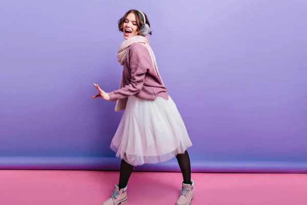 Cheerful girl with short haircut wears black pantyhose funny posing in studio. Glad european lady in long skirt and knitted scarf fooling around on purple background..