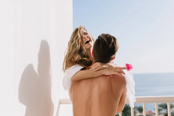 Laughing girl in white blouse having fun on terrace spending summer vacation with her boyfriend. Beautiful happy couple hugging on balcony with sea views on honeymoon on exotic resort.