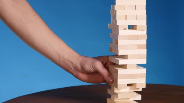 Mãos de Jovem joga jenga no fundo azul, close-up. Um homem constrói uma torre de blocos enquanto joga jenga — Vídeo de Stock