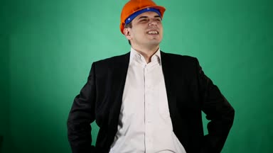 Bir portakal inşaat kask ve takım elbise çekici bir adam kameraya benziyor. Genç bir işçi hareketleri elleri ile yeşil chroma anahtar arka plan üzerinde gösterir..