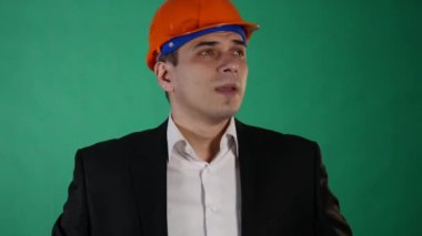 Bir portakal inşaat kask ve takım elbise çekici bir adam kameraya benziyor. Genç bir işçi hareketleri elleri ile yeşil chroma anahtar arka plan üzerinde gösterir.