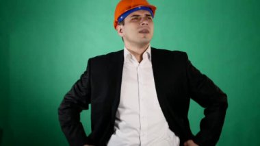 Bir portakal inşaat kask ve takım elbise çekici bir adam kameraya benziyor. Genç bir işçi hareketleri elleri ile yeşil chroma anahtar arka plan üzerinde gösterir.