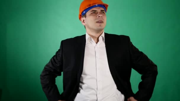 An attractive man in a orange construction helmet and suit looks at the camera. A young worker shows gestures with his hands on green chroma key background — Stock Video