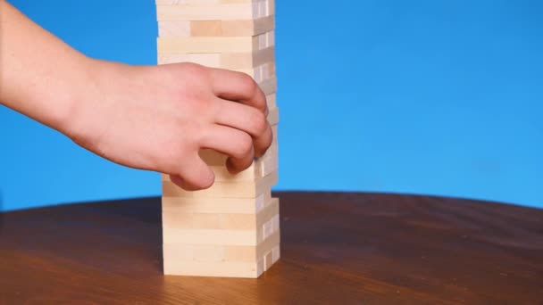 Manos de Joven juega jenga sobre fondo azul, de cerca. Un hombre construye una torre de bloques mientras juega jenga — Vídeos de Stock