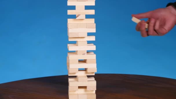 Mãos de Jovem joga jenga no fundo azul, close-up. Um homem constrói uma torre de blocos enquanto joga jenga — Vídeo de Stock