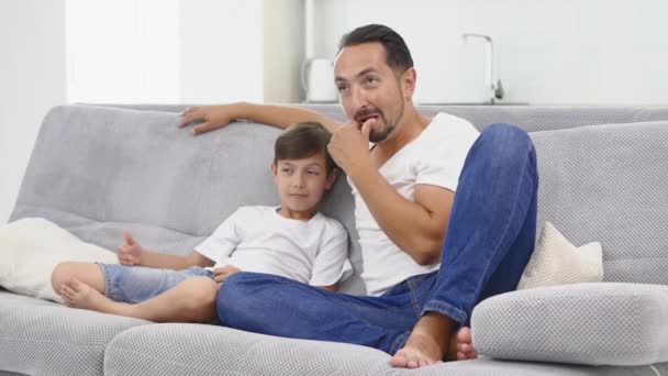 A ver televisão deitada no sofá. menino e seu pai estão assistindo TV — Vídeo de Stock