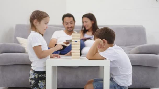 Šťastné děti bavit hraním Jenga v obývacím pokoji. rodiče budou číst časopis na pozadí — Stock video