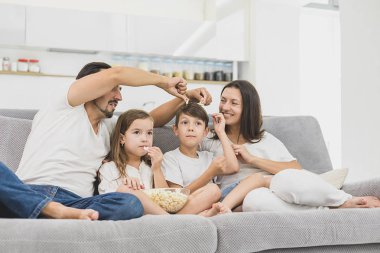 Güzel, genç ebeveynler ve çocukları televizyon izliyor, patlamış mısır yiyor ve evde kanepede otururken gülümsüyorlar..