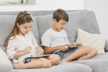 Evde kanepede oturan ve pc tablet ile birlikte oynayan mutlu kardeşler