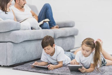 Aile, çocuk, teknoloji ve ev kavramı - kardeşi ile tablet pc bilgisayar ve veliler arkadaki kitaplarla bir kız kardeşle