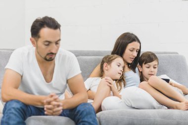 Kanepede oturan ve evde cep telefonuyla iki çocuk ile anne. Baban üzgün