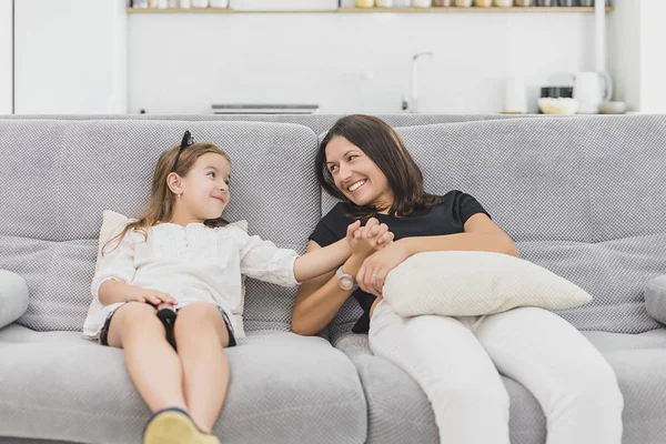 Мати і дочка веселяться у вітальні, сидячи на сірому дивані. мати і дочка тримаються за руки — стокове фото