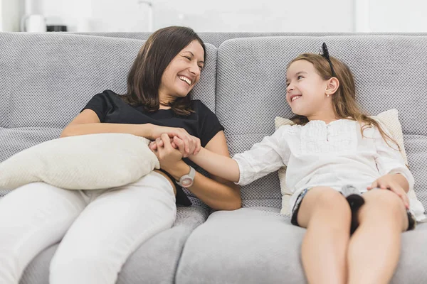 Madre e hija se están divirtiendo mucho en la sala de estar sentadas en el sofá gris. madre e hija cogidas de la mano — Foto de Stock