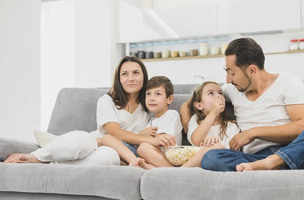 Feliz familia joven viendo la televisión con padres jóvenes y atractivos sentados con sus dos hijos en un sofá en la sala de estar —  Fotos de Stock