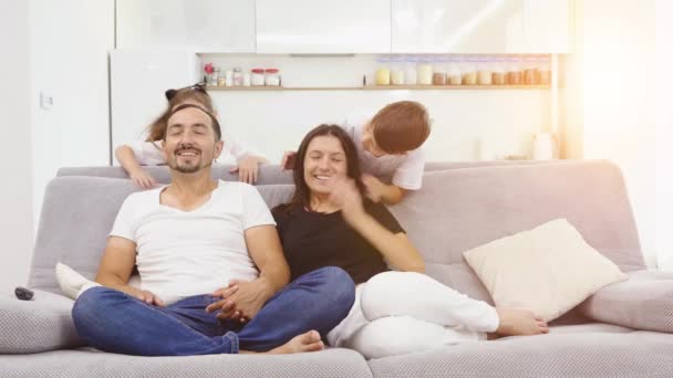 Счастливая семья сидит на диване и смотрит телевизор. родители смотрят телевизор, а их дети подкрадываются сзади — стоковое видео