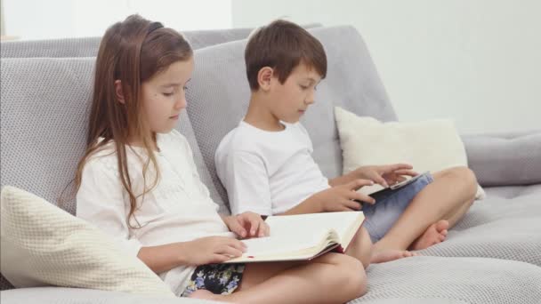 Little cute boy play tablet while his sister reads a book on sofa at home. Modern communication and gadget addiction concept. Side view — Stock Video