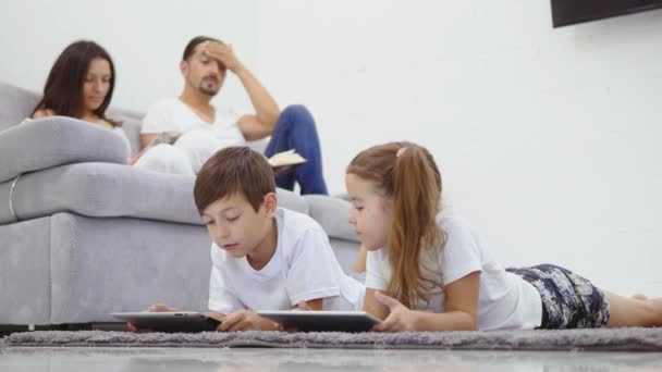 Familj, barn, teknik och hem koncept - bror med en syster med tablet pc-dator och föräldrar på baksidan med böcker. underifrån — Stockvideo