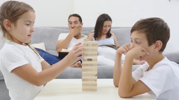 Fröhliche Kinder, die Spaß beim Jenga-Spielen im Wohnzimmer haben. Eltern gähnen im Hintergrund — Stockvideo