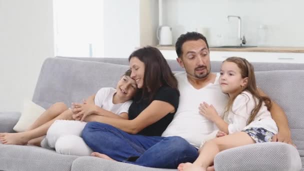 Ocio juntos. La familia feliz de cuatro está disfrutando en casa. Los niños pequeños y lindos están con juguetes, los padres están en el sofá, abrazándose, viendo dibujos animados — Vídeo de stock