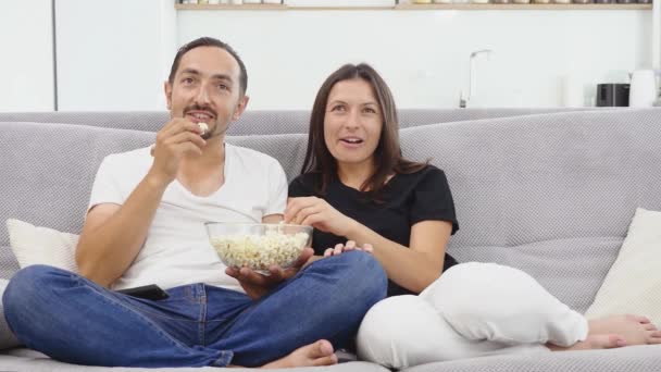 Счастливая расслабленная пара сидит на диване смотреть вместе фильм по телевизору и есть попкорн — стоковое видео
