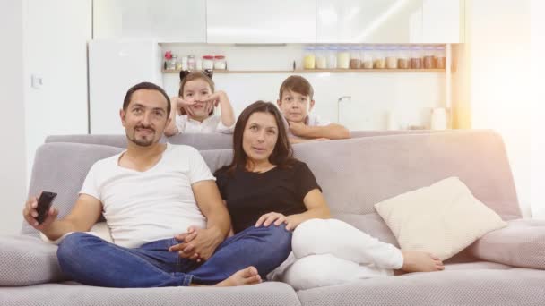 Feliz familia sentada en el sofá viendo la televisión. los padres están sentados en el sofá y los niños están detrás — Vídeos de Stock