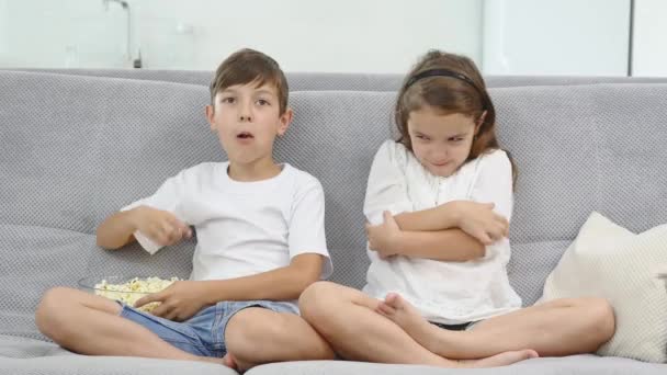 Милі діти їдять попкорн під час перегляду телевізора вдома. маленька дівчинка ображається, тому що брат не дає їй попкорн — стокове відео