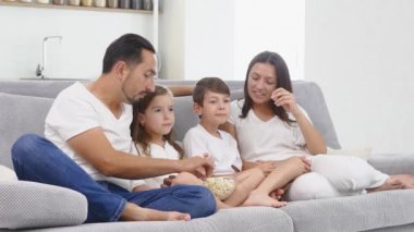 Güzel, genç ebeveynler ve çocukları televizyon izliyor, patlamış mısır yiyor ve evde kanepede otururken gülümsüyorlar.