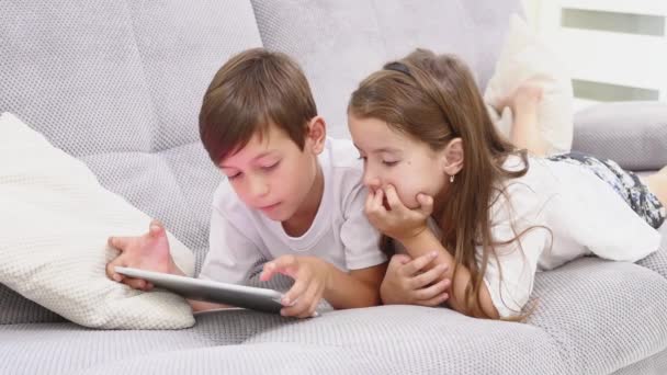 Dvě malé děti hrát hry a vzdělávací aplikace online pomocí počítače tablet pc, těší a s fun ležet na gauči doma během slunečného dne — Stock video