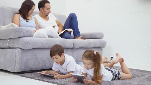 Familj, barn, teknik och hem koncept - bror med en syster med tablet pc-dator och föräldrar på baksidan med böcker — Stockvideo