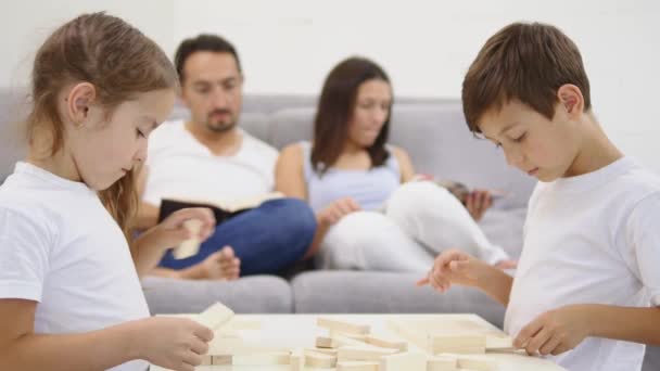 Gelukkige kinderen plezier spelen Jenga in de woonkamer. ouders lezen boeken op de achtergrond — Stockvideo