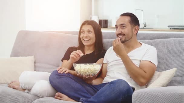 Um casal a ver televisão. Sentam-se num sofá na sua acolhedora sala de estar e comem pipocas — Vídeo de Stock