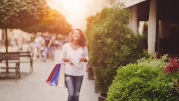 Egy fiatal nő, bevásárló táskák megy az utcán, és napszemüveget visel. Sunshine háttér — Stock videók
