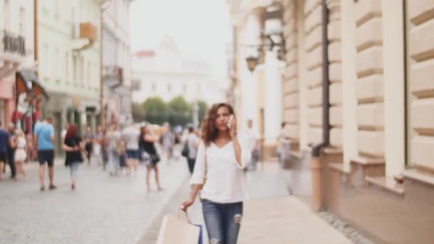 Hermosa joven con bolsas de compras usando su teléfono inteligente. Linda chica caminando en una calle de la ciudad después de ir de compras y hablar por teléfono — Vídeos de Stock