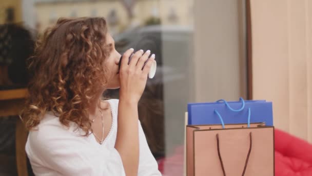 Elegante jongedame met koffie en boodschappentassen rusten in de buurt van venster op straat — Stockvideo