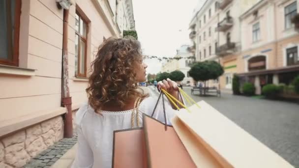 Bakifrån av ung kvinna med shoppingkassar njuter i shopping, tjej har roligt med sina inköp i city. Konsumtion och livsstil koncept. Slow motion — Stockvideo