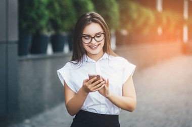 Açık havada gülümseyen bir güzel şehirde kadın bir klasik finansal holding bir akıllı telefon kullanarak ofis binaları ile sokak portresi. Profesyonel erkek güneşli dış teknolojisini kullanarak
