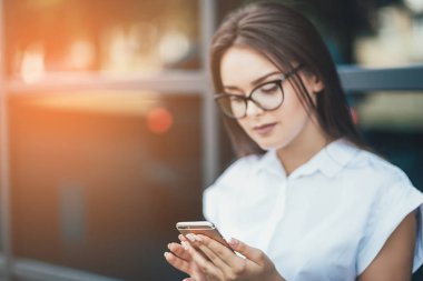 Beyaz gömlek giymiş ve kopma şehirde yürürken modern akıllı telefon kullanarak güzel genç bir işkadını. Güneş arka plan