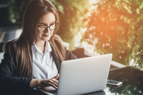 Wanita muda yang bahagia duduk di kafe dan menggunakan laptop. Latar belakang sinar matahari — Stok Foto