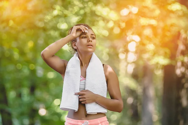 Young attractive woman relaxing from doing exercise in a park city and drying her forehead sweat with a white towel during a sunny morning — Stock Photo, Image