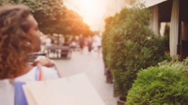 Back view of attractive curly brunette woman walking down with shopping bags in the city-street or alley, turns to camera and gives a beautiful smile. Sunshine on the background — Stock Video