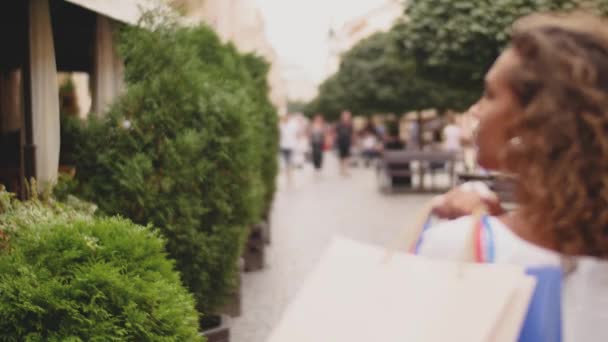 Вид сзади стильной молодой женщины с сумками для покупок, идущей по улице — стоковое видео