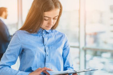The secretary with a folder and documents is in the office. During this she is on the background of her colleagues.