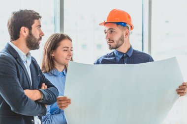 Mimar bir turuncu kutu ofiste iş eşleri yanında duruyor. Bu sırada o ortakları gösterilen bir iş planı tutar.