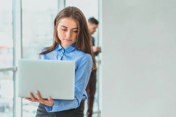 The secretary is standing in the office holding the laptop in his hands. During this she is on the background of her colleagues. — Stock Photo, Image