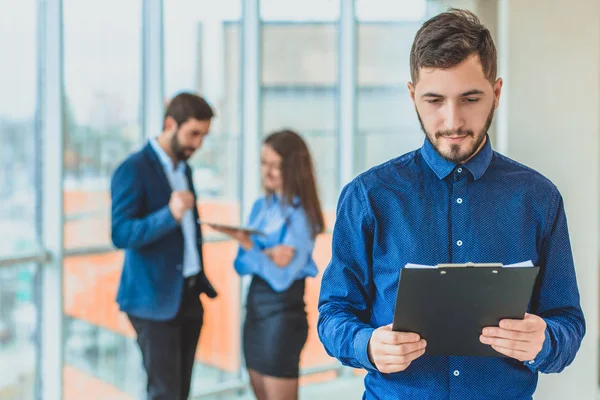 Biznesmenem stoi w biurze gospodarstwa folder w jego ręce. W tym czasie on stoi na tle kolegów, którzy pracują na tablecie. — Zdjęcie stockowe