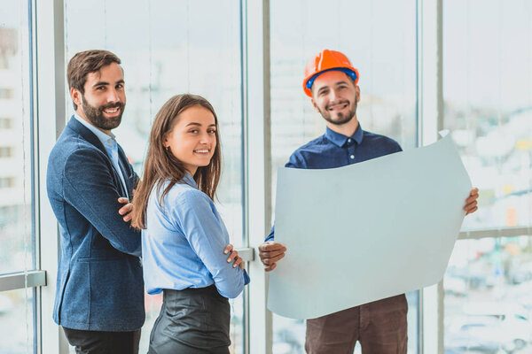 An architect in an orange box office is standing next to their business partners. During this, he keeps a business plan showing his partners.