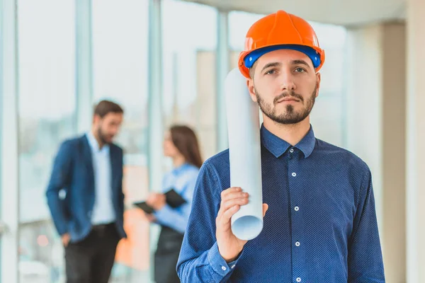 The architect is in a cask of orange color. In the office against the background of their colleagues.